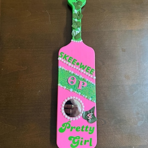 Puede incluir: Una paleta rosa con detalles y texto en verde. La paleta presenta las palabras "Skee Wee" y "Pretty Girl" en verde, junto con las letras "OP" y un espejo. Adornada con perlas y hojas.