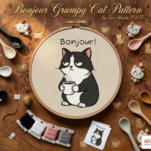 May include: A cross-stitch pattern featuring a grumpy black and white cat holding a coffee cup, with the word "Bonjour!" above it. The image includes embroidery supplies, cat-shaped pins, and spoons, all arranged on a wooden surface.