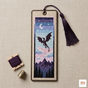 May include: A handmade bookmark featuring a cross-stitch design of a dragon in flight against a night sky with a crescent moon and stars. The bookmark has a dark purple tassel and a metal frame. A spool of purple thread and a thimble are next to the bookmark.