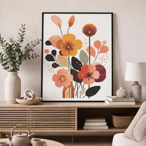 May include: A framed floral cross-stitch pattern featuring a bouquet of stylized flowers in shades of orange, peach, and burgundy. The artwork includes black accents and is set against a white background. The image is displayed in a living room setting.