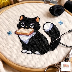Op de afbeelding: Een kruissteekproject in een houten borduurring met een zwarte kat met witte pootjes, een witte borstvlek en een wit puntje aan de staart. De kat houdt een beige rechthoekig object in de bek. Kleine blauwe kruissteeksterren omringen de kat.