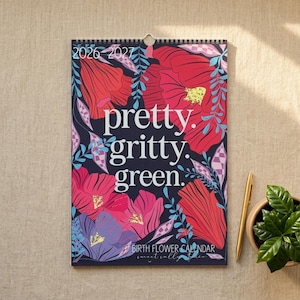 Könnte beinhalten: Wandkalender 2026-2027 mit floralem Design und den Worten "pretty. gritty. green." in Weiß. Der Kalender hat einen dunkelblauen Hintergrund mit roten, rosa und lila Blumen. Ein goldener Stift und eine kleine Topfpflanze liegen neben dem Kalender.