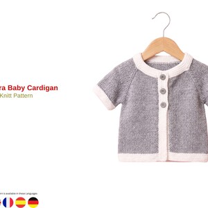 May include: A short-sleeved baby cardigan in heather gray with white trim on the collar, cuffs, and hem. It features three small gray buttons down the front and is displayed on a wooden hanger. The text reads Mundra Baby Cardigan Knit Pattern.