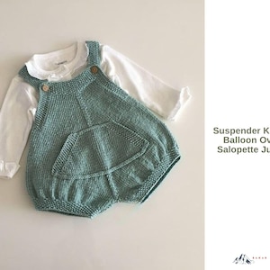 Puede incluir: Un peto de bebé tejido en verde azulado con un bolsillo delantero grande y botones de madera, superpuesto a una camisa blanca de manga larga. El peto es de estilo globo y la camisa tiene un cuello con volantes.