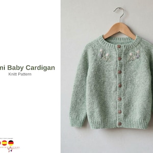 May include: A light green baby cardigan with a round neck and long sleeves, hanging on a wooden hanger. The knitted sweater features small floral embroidery near the neckline and is fastened with brown buttons. The text "Kremi Baby Cardigan Knitt Pattern" is visible.