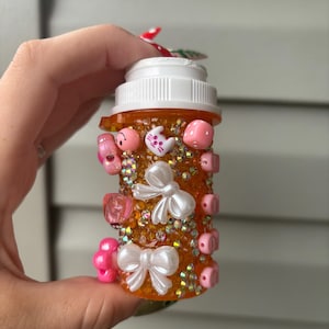 May include: An orange pill bottle decorated with pink and white embellishments. The bottle is adorned with small pink beads, white bows, and various pink charms, including a cat and a mushroom. The bottle is filled with iridescent glitter.