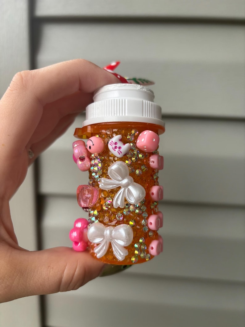 May include: An orange pill bottle decorated with pink and white embellishments. The bottle is adorned with small pink beads, white bows, and various pink charms, including a cat and a mushroom. The bottle is filled with iridescent glitter.