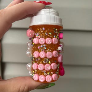 May include: An orange pill bottle decorated with iridescent glitter and pink beads. The bottle has the words "TAKE YOUR DAMN MEDS" spelled out in pink letter beads. The bottle is held by a person with long, red nails.