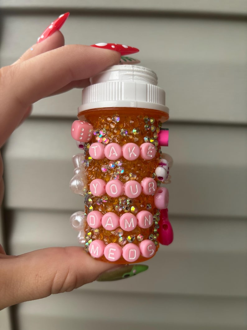 May include: An orange pill bottle decorated with iridescent glitter and pink beads. The bottle has the words "TAKE YOUR DAMN MEDS" spelled out in pink letter beads. The bottle is held by a person with long, red nails.
