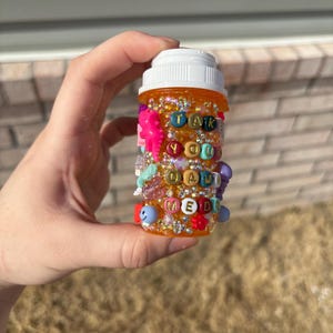 May include: An orange pill bottle decorated with colorful beads and the words "TAKE YOUR DAMN MEDS". The bottle is adorned with various charms, including a pink seahorse and smiley face beads.