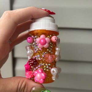 May include: An orange pill bottle decorated with pink flowers, beads, and rhinestones. The bottle has a white cap and is embellished with various charms, including dice and heart-shaped beads. The bottle is held by a person with long, decorated nails.
