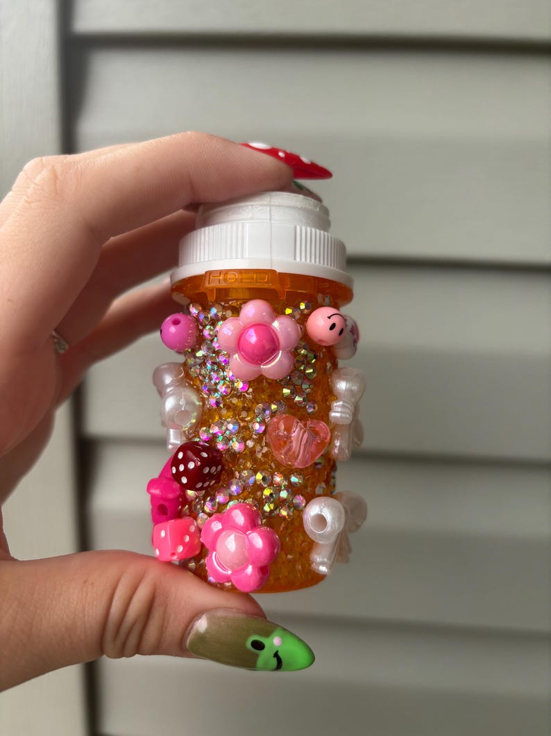 May include: An orange pill bottle decorated with pink flowers, beads, and rhinestones. The bottle has a white cap and is embellished with various charms, including dice and heart-shaped beads. The bottle is held by a person with long, decorated nails.