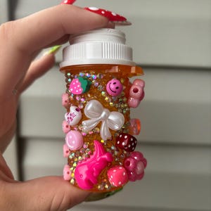 May include: A decorated orange pill bottle adorned with various charms, including a white bow, pink smiley faces, strawberries, and dice. The bottle is covered in small, sparkling embellishments.