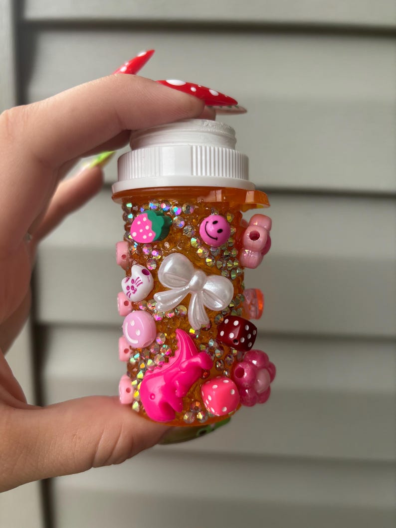 May include: A decorated orange pill bottle adorned with various charms, including a white bow, pink smiley faces, strawberries, and dice. The bottle is covered in small, sparkling embellishments.