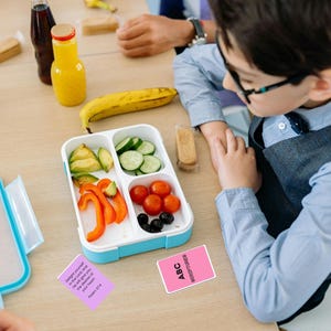 May include: A child's lunchbox filled with sliced avocado, cucumbers, bell peppers, cherry tomatoes, and olives. A yellow bottle of juice and a banana are also on the table. Two cards with text are visible.