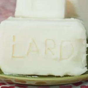 May include: A block of white lard with the word "LARD" carved into the surface, on a green plate. Another block of lard is visible behind it. The plate is on a surface patterned with red hearts.