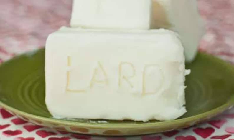 May include: A block of white lard with the word "LARD" carved into the surface, on a green plate. Another block of lard is visible behind it. The plate is on a surface patterned with red hearts.