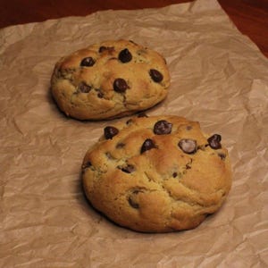 Könnte beinhalten: Zwei Schokoladen-Chip-Cookies auf braunem Backpapier. Die Kekse sind goldbraun mit sichtbaren Schokostückchen. Das Papier hat eine zerknitterte Textur. Ein einfaches, einladendes Bild.