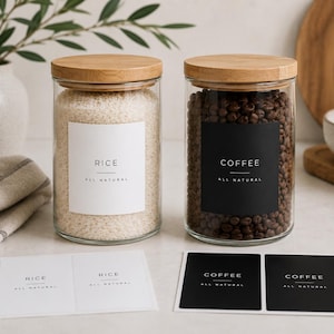 以下が含まれることがあります： 明るい木製の蓋が付いた2つの透明な円筒形のガラス製保存瓶。1つは白米が入っており、RICE ALL NATURALと書かれた白いラベルが付いています。もう1つはコーヒー豆が入っており、COFFEE ALL NATURALと書かれた黒いラベルが付いています。