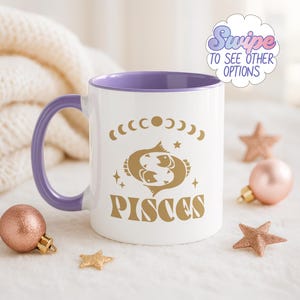 Op de afbeelding: Witte keramische mok met een paarse binnenkant en handvat. De mok heeft een gouden Vissen-sterrenbeeldontwerp met manen, sterren en het woord "PISCES". De afbeelding bevat decoratieve sterren en een kerstbal.