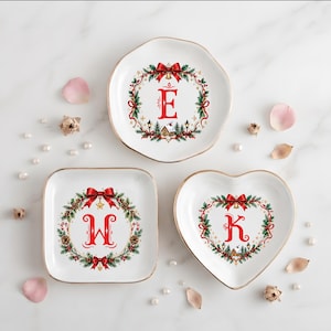 Könnte beinhalten: Drei weiße Keramikschalen mit goldfarbenen Rändern, verziert mit weihnachtlichen Kranzmotiven in Rot und Grün. Die Schalen zeigen die Buchstaben E, W und K in Rot, umgeben von Tannenzweigen, roten Schleifen und kleinen Häusern.