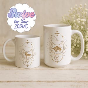 Op de afbeelding: Twee witte keramische mokken met gouden dierenriemdesigns. De mokken bevatten illustraties van sterrenbeelden en dierenriemsymbolen. Eén mok is kleiner dan de andere. De afbeelding bevat de tekst "Swipe For Your ZODIAC".