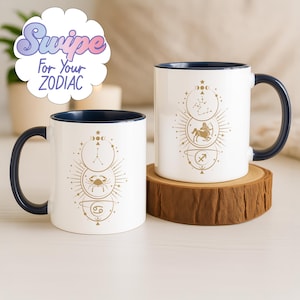 Op de afbeelding: Twee witte keramische mokken met marineblauwe binnenkant en handvatten. Elke mok heeft een gouden dierenriemdesign met sterrenbeelden en symbolen. Eén mok toont de tekst "Swipe For Your Zodiac" in een kleurrijk wolkontwerp.
