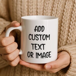 Può includere: Tazza in ceramica bianca con manico curvo. La tazza presenta il testo "ADD CUSTOM TEXT OR IMAGE" in grassetto nero. La tazza è tenuta da una persona che indossa un maglione beige.