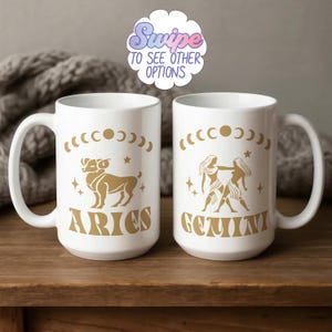 Op de afbeelding: Twee witte keramische mokken met gouden dierenriemdesigns. Eén mok toont een Ram, de andere een Tweelingen-illustratie. Beide mokken hebben de woorden "ARIES" en "GEMINI" in goud. De tekst "Swipe to see other options" staat bovenaan.