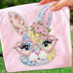 Op de afbeelding: Een roze stof met een geborduurd konijnengezicht. Het konijn heeft hartvormige brillen, lange wimpers en een roze neus. De oren van het konijn zijn versierd met bloemmotieven en parels. Een tros roze rozen zit bovenop het hoofd van het konijn. Het ontwerp staat op een roze achtergrond.
