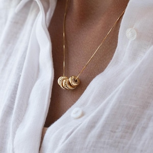 May include: Gold-colored necklace with multiple circular pendants. Each pendant has a unique design, with some featuring small, sparkling stones. The necklace is worn against a white shirt.