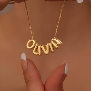 Könnte beinhalten: Eine goldfarbene Halskette mit dem Namen "OLIVIA" in großen, blasenartigen Buchstaben. Die Halskette hängt an einer zarten Kette. Der Hintergrund ist verschwommen und warm getönt.