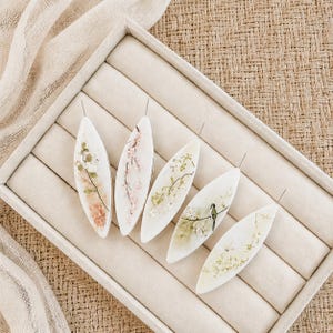 May include: Five elongated, leaf-shaped white resin pendants, each featuring embedded dried flowers and foliage. One pendant includes a small bird figure. Each piece has a thin metal pin extending from the top, arranged in a beige jewelry display tray.