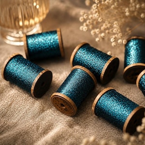 May include: Several wooden spools wrapped in shimmering teal metallic thread. Each spool features a cylindrical wooden core with flat circular ends, showcasing a dense, reflective texture that catches the light across the vibrant blue-green surface.