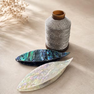 Puede incluir: Un carrete de hilo plateado metálico colocado detrás de dos pinzas para el cabello en forma de hoja. Una pinza presenta un patrón de concha de abulón iridiscente con tonos verdes y azules, mientras que la otra muestra un acabado de concha blanco nacarado.