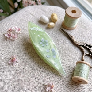 May include: A light green, leaf-shaped tatting shuttle with a pearlescent finish, decorated with white floral and butterfly motifs. It is surrounded by two wooden spools of light green thread, vintage-style metal scissors, and small pink flowers.