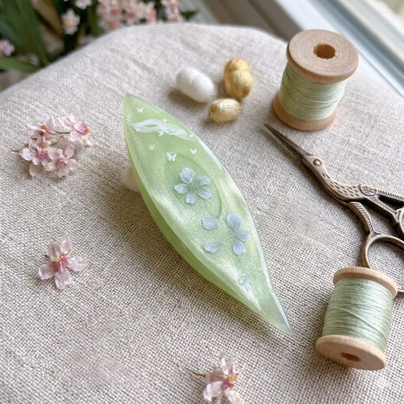 May include: A light green, leaf-shaped tatting shuttle with a pearlescent finish, decorated with white floral and butterfly motifs. It is surrounded by two wooden spools of light green thread, vintage-style metal scissors, and small pink flowers.