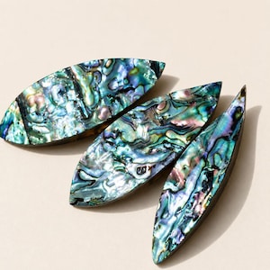 Könnte beinhalten: Drei marquiseförmige Abalone-Muschel-Cabochons mit einer polierten, irisierenden Oberfläche mit wirbelnden Mustern in tiefem Blau, leuchtendem Grün sowie Nuancen von Violett und Rosa, die einen natürlichen, perlmuttartigen Glanz aufweisen.