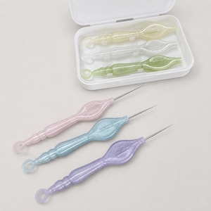 Peut inclure: Un ensemble de six outils de loisirs créatifs avec des manches translucides et pailletés aux couleurs pastel, notamment jaune, transparent, vert, rose, bleu et violet. Trois outils sont rangés dans un boîtier rectangulaire blanc en plastique.