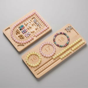 May include: Two light-colored wooden bead design boards with carved channels and numbered markings. One board features a U-shaped channel filled with pink beads, while the other displays three circular bracelets and a straight channel with yellow beads.