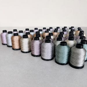 Puede incluir: Una colección de numerosos carretes de hilo de bordar metálico dispuestos en formación triangular. Cada carrete presenta una base y una parte superior de plástico negro, con colores de hilo que incluyen tonos plata, oro, bronce, lavanda y verde azulado.