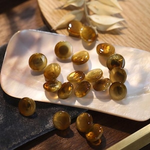 Peut inclure: Une collection de petits boutons ronds de couleur ambre avec une finition lisse et brillante. Les boutons sont dispersés sur un plat rectangulaire en coquillage blanc irisé, certains reposant sur le tissu sombre sous le plat.