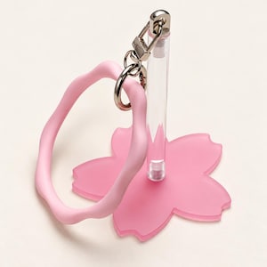 Puede incluir: Una pulsera de silicona ondulada de color rosa claro unida a un cierre de llavero de metal plateado. El llavero está montado sobre una varilla vertical transparente que se apoya en una base plana rosa claro mate con forma de flor de cerezo.