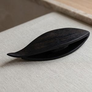 Op de afbeelding: Een bladvormig weefspoeltje van donkerbruin hout met een gladde, gepolijste afwerking. Het object heeft een puntig uiteinde en een holle, langwerpige opening in het midden, waarbij de natuurlijke houtnerf zichtbaar is over de hele vorm.