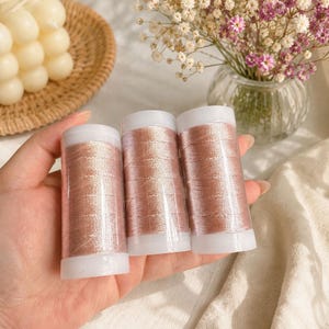 Puede incluir: Tres carretes cilíndricos de hilo de coser color oro rosa metálico. Cada carrete cuenta con una tapa de plástico blanca en la parte superior e inferior, con un hilo brillante y reflectante enrollado firmemente alrededor del centro.