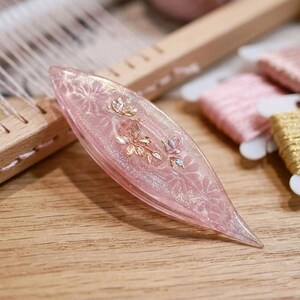 May include: A translucent pink tatting shuttle with a pointed, almond-shaped design. The surface features embedded gold-colored floral accents and a subtle glitter finish. The shuttle is crafted from a smooth, glossy material with a tapered profile.