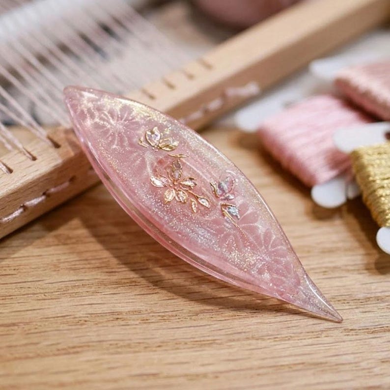 May include: A translucent pink tatting shuttle with a pointed, almond-shaped design. The surface features embedded gold-colored floral accents and a subtle glitter finish. The shuttle is crafted from a smooth, glossy material with a tapered profile.