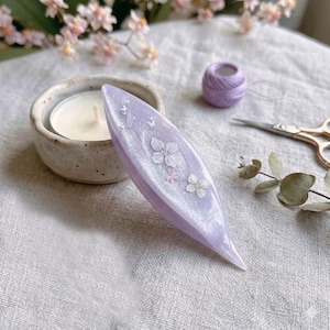 May include: A lavender tatting shuttle with a pearlescent finish, featuring embedded white floral decals and small butterfly motifs. The shuttle has a pointed, elongated oval shape and is positioned near a small candle, thread, and scissors.
