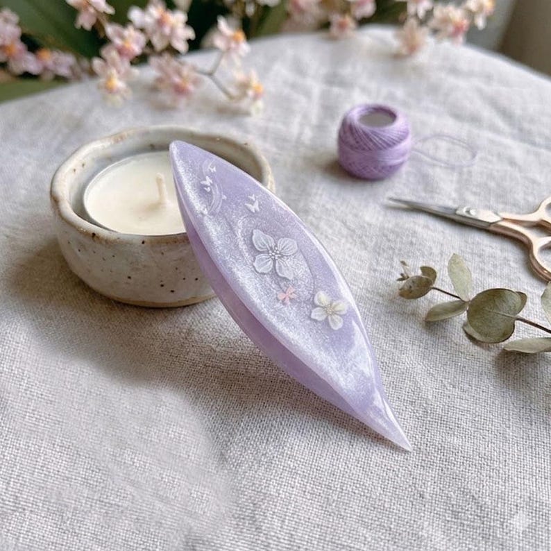 May include: A lavender tatting shuttle with a pearlescent finish, featuring embedded white floral decals and small butterfly motifs. The shuttle has a pointed, elongated oval shape and is positioned near a small candle, thread, and scissors.
