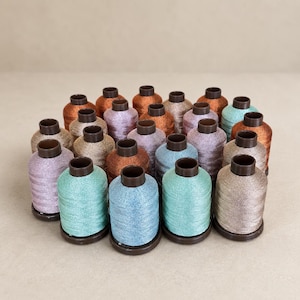 May include: A collection of twenty-four cylindrical spools of metallic embroidery thread arranged in a cluster. The spools feature a variety of shimmering colors including shades of copper, bronze, light blue, mint green, and soft lavender.